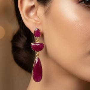 Könnte beinhalten: Goldfarbene Ohrhänger mit drei rubinroten Edelsteinen. Der obere Stein ist oval, der mittlere halbkreisförmig und der untere tropfenförmig. Die Ohrringe werden an einem Ohr getragen.