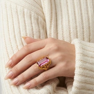May include: A gold ring featuring a large, rectangular, faceted pink gemstone set in an ornate, filigree-style metal band. The setting has a decorative, textured gold finish that surrounds the base of the vibrant, translucent pink stone.