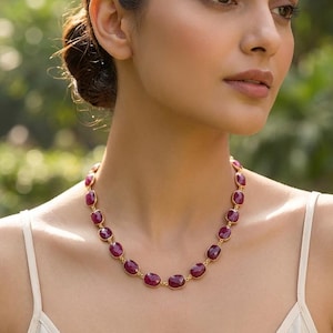 May include: A necklace featuring a series of oval-shaped, deep magenta-red gemstones set in gold-toned metal bezels. The stones are linked together in a continuous chain, creating a uniform pattern that rests around the base of the neck.