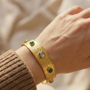 Könnte beinhalten: Ein goldfarbenes Armband mit einer texturierten, körnigen Oberfläche. Das Armband ist mit mehreren kleinen, runden, in Fassungen gesetzten Cabochon-Steinen in abwechselnden Farbtönen von tiefem Grün und weichem, durchscheinendem Grau verziert.