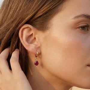 Puede incluir: Un pendiente de aro dorado con un pequeño colgante de piedra preciosa de color rojo intenso en forma de lágrima. El pendiente está sujeto al lóbulo de la oreja, mostrando un acabado metálico pulido en el aro y una superficie lisa en la piedra.