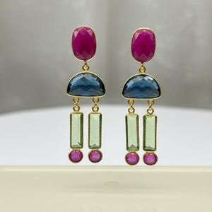 Puede incluir: Un par de pendientes dorados con un diseño geométrico. Cada pendiente presenta una piedra ovalada magenta en la parte superior, una piedra azul en forma de semicírculo, dos piedras rectangulares verdes y dos piedras redondas magenta. Los pendientes se exhiben sobre una superficie transparente.