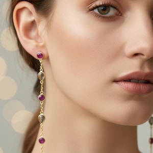 Könnte beinhalten: Goldfarbene Ohrhänger mit abwechselnd rubinroten und rauchgrauen Edelsteinen. Die Ohrringe haben tropfenförmige und runde Edelsteine, die an einem kleinen Stecker befestigt sind und einen zarten, hängenden Effekt erzeugen.