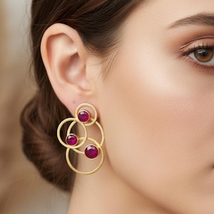 Peut inclure: Boucles d'oreilles dorées composées de cercles interconnectés, chacun orné d'une pierre précieuse rouge ronde et vibrante. Le design est moderne et abstrait, mélangeant formes géométriques et couleurs vives.