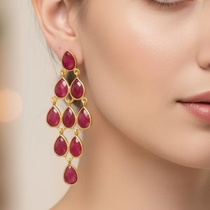 Può includere: Orecchini a lampadario dorati con pietre preziose rosse a forma di goccia. Gli orecchini hanno un design a strati, con ogni pietra incastonata in una lunetta dorata. Un gioiello appariscente per occasioni speciali.