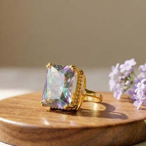 Peut inclure: Une bague dorée ornée d'une grande pierre précieuse rectangulaire iridescente avec une finition prismatique multicolore. Le sertissage présente un bord perlé décoratif autour de la pierre, et l'anneau est en métal doré lisse.