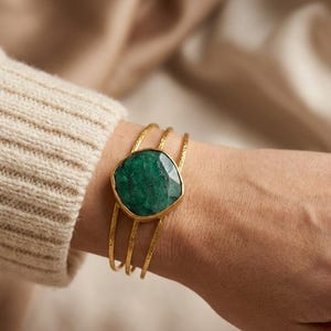 Op de afbeelding: Een goudkleurige armband met drie dunne, getextureerde banden en een grote, gefacetteerde, vierkante groene edelsteen in een goudkleurige zetting. De armband wordt om de pols gedragen en toont het contrast tussen de gouden afwerking en de diepgroene steen.