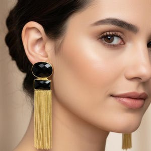 Può includere: Orecchini color oro con accenti di pietre nere e frange di catena pendenti. Gli orecchini presentano un design ovale e rettangolare in pietra nera. La frangia pende sotto la pietra rettangolare, creando un look lungo ed elegante.