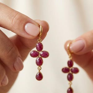 May include: A pair of gold drop earrings featuring a cross-shaped arrangement of four oval-cut deep pink rubies, with a single pear-cut ruby dangling at the bottom of each earring. The stones are set in gold bezels and attached to gold hoop findings.