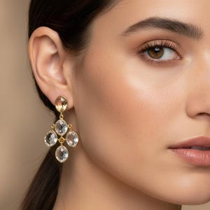 May include: Gold-toned chandelier earrings with clear, teardrop-shaped crystals. The earrings have a cluster design, with a single, smaller crystal at the top and four larger crystals below. The earrings are a statement piece of jewelry.