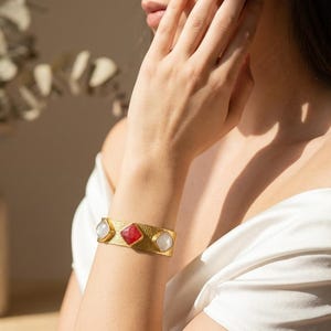 Puede incluir: Un brazalete tipo puño de tono dorado con una superficie texturizada y tres piedras de corte cuadrado. La piedra central es de un rojo vibrante, flanqueada por dos piedras blancas translúcidas a cada lado. El brazalete tiene un diseño ancho.