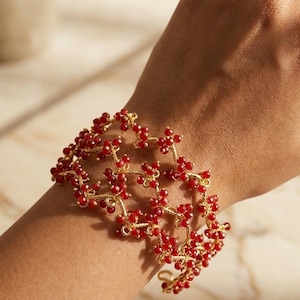 May include: A gold-toned cuff bracelet featuring a delicate, interconnected wire structure adorned with numerous small, round, vibrant red beads. The design creates a repeating geometric pattern that wraps around the wrist, showcasing a textured metallic finish.