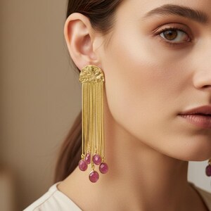 May include: Gold-coloured earrings featuring a textured, semi-circular top and hanging chains. Each chain has a small, oval, pink gemstone. These earrings are a statement piece, adding elegance and glamour.