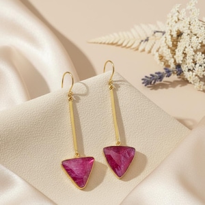 May include: A pair of gold-colored dangle earrings. Each earring features a long, thin bar suspended from a hook, with a triangular, magenta-colored gemstone at the bottom. The earrings are displayed on a cream-colored surface with a satin texture.