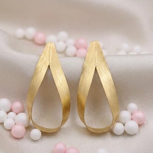 May include: Two gold-coloured teardrop earrings with a brushed metal finish. The earrings are presented on a satin surface, with scattered pink and white beads.