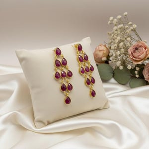 May include: A pair of gold-toned chandelier earrings with cascading, teardrop-shaped, deep red gemstones. The earrings are displayed on a white pillow against a backdrop of white satin fabric and flowers.