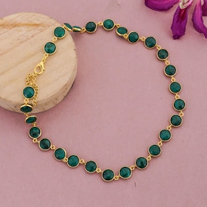 May include: A gold-colored necklace featuring a series of round, emerald-green gemstones. The gemstones are evenly spaced and connected by delicate gold links. The necklace is displayed on a light pink surface.