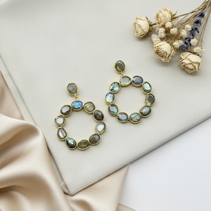 May include: A pair of gold-toned hoop earrings. Each earring features a circular design adorned with iridescent, oval-shaped stones in shades of gray, blue, and green. The earrings rest on a light gray surface, with dried flowers in the background.