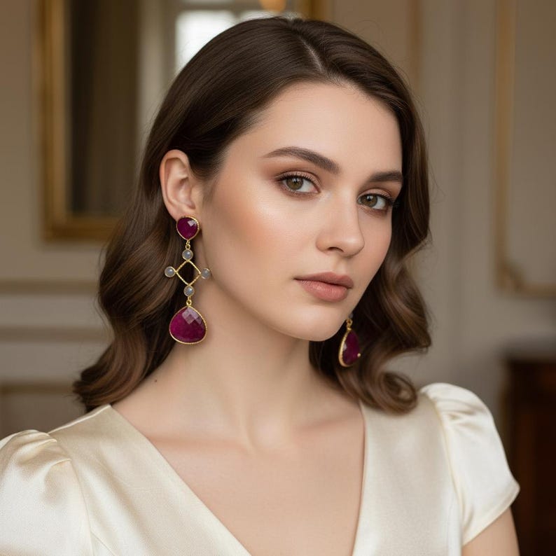 May include: Gold-coloured earrings with large, faceted, deep red gemstones. The earrings feature a geometric design with smaller grey stones and a teardrop-shaped ruby. The woman in the image is wearing a cream-coloured dress.