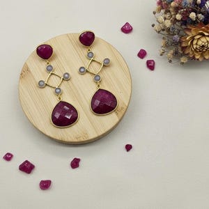 May include: Gold-coloured earrings featuring large, faceted, ruby-red teardrop-shaped stones. The earrings have a geometric design with smaller grey stones. They are displayed on a wooden surface with scattered ruby-red gems.