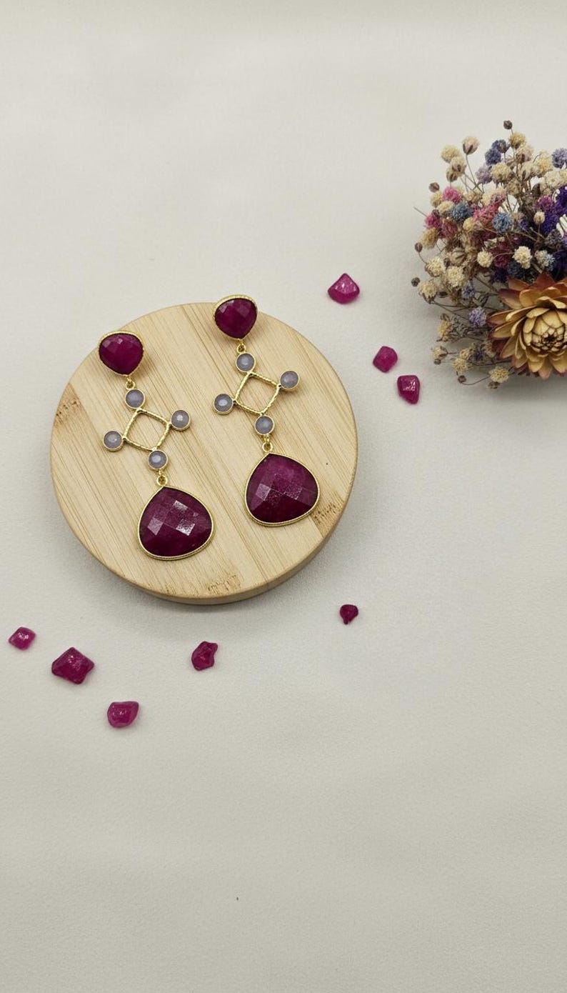 May include: Gold-coloured earrings featuring large, faceted, ruby-red teardrop-shaped stones. The earrings have a geometric design with smaller grey stones. They are displayed on a wooden surface with scattered ruby-red gems.