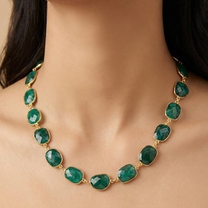 May include: A gold necklace with large, emerald-green gemstones. The oval-shaped gemstones are linked by gold-coloured metal. The necklace is displayed on a person's neck, highlighting its design and colour. The gemstones are approximately 1.5 cm in length.