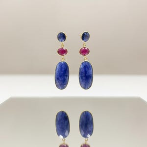 Può includere: Un paio di orecchini pendenti color oro. Ogni orecchino presenta una piccola gemma blu in alto, una gemma rossa di medie dimensioni al centro e una grande gemma blu di forma ovale in basso. Gli orecchini sono esposti su una superficie riflettente.