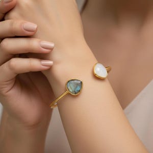 Op de afbeelding: Een goudkleurige manchetarmband met twee edelsteenaccenten. Eén accent is een peervormige, iriserende blauwgroene steen, de andere is een witte, driehoekige steen. De armband wordt om een pols gedragen en toont het ontwerp en de kleur van het sieraad.