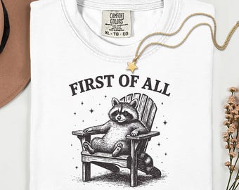 May include: White t-shirt with a raccoon illustration sitting in a wooden chair. The text "FIRST OF ALL I'M A DELIGHT" is printed below the raccoon. A gold star pendant necklace and a brown hat are also visible.