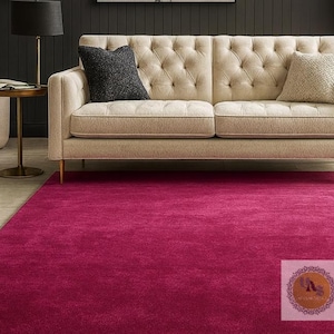 May include: A magenta-colored area rug covers the floor in front of a beige tufted sofa with two throw pillows. A black lamp with a black shade sits on a gold side table. The rug is a solid color and appears soft.