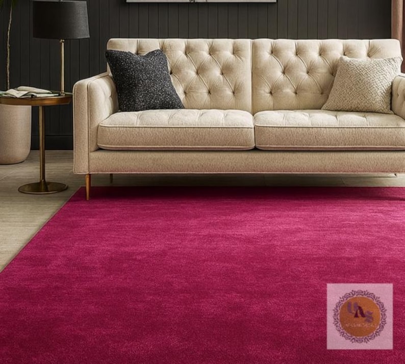 May include: A magenta-colored area rug covers the floor in front of a beige tufted sofa with two throw pillows. A black lamp with a black shade sits on a gold side table. The rug is a solid color and appears soft.
