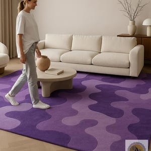 May include: A purple area rug with a wavy pattern in shades of violet and lavender. The rug is placed in a living room setting with a cream-colored sofa, a round coffee table, and a person walking across the rug.
