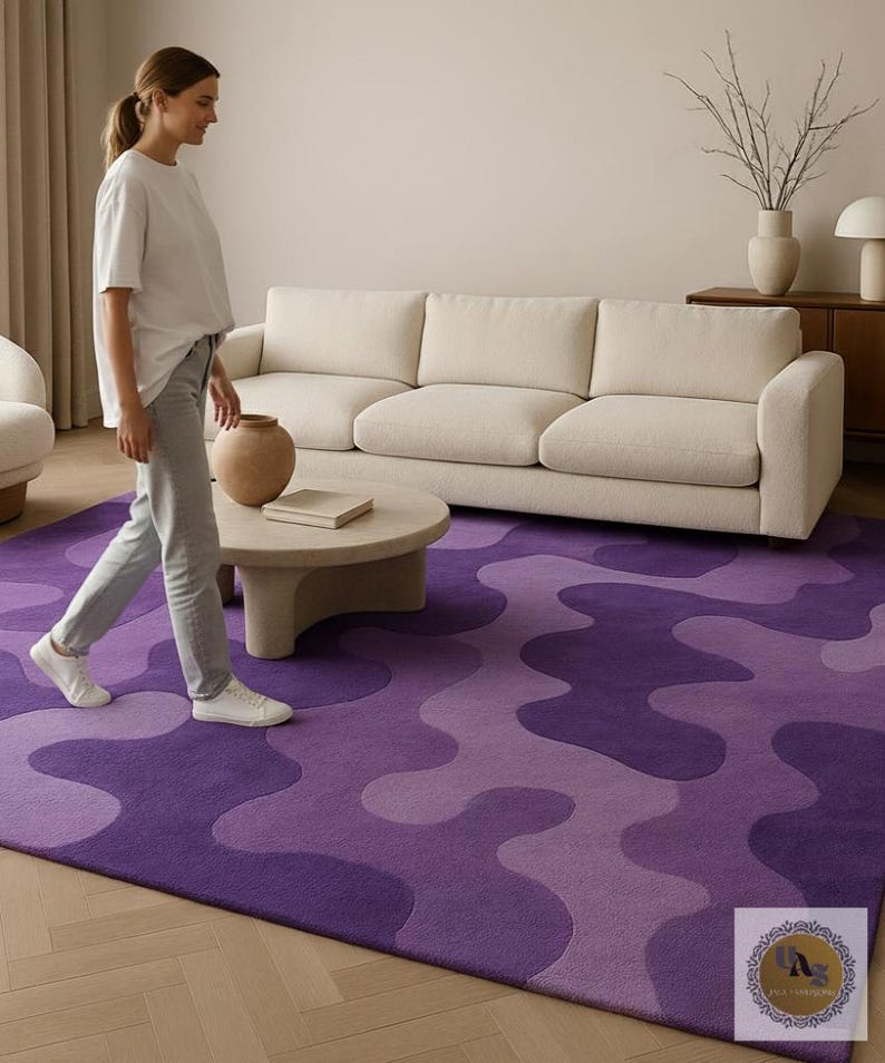 May include: A purple area rug with a wavy pattern in shades of violet and lavender. The rug is placed in a living room setting with a cream-colored sofa, a round coffee table, and a person walking across the rug.