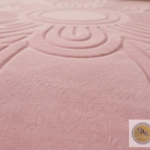 Puede incluir: Primer plano de una alfombra de color rosa empolvado con un dise&ntilde;o floral circular en relieve. La alfombra tiene una superficie suave y texturizada y un patr&oacute;n detallado. El dise&ntilde;o est&aacute; centrado e irradia hacia afuera.
