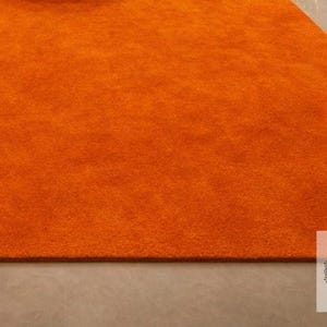 May include: A section of an orange rug with a soft, textured surface. The rug is a solid color and appears to be made of a plush material. The rug is on a light gray surface. The rug is a vibrant accent piece.