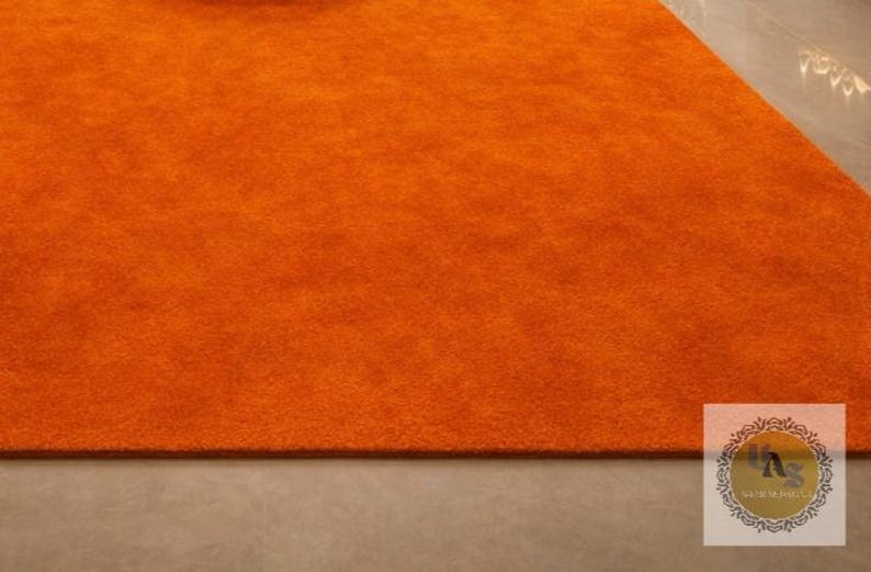 May include: A section of an orange rug with a soft, textured surface. The rug is a solid color and appears to be made of a plush material. The rug is on a light gray surface. The rug is a vibrant accent piece.