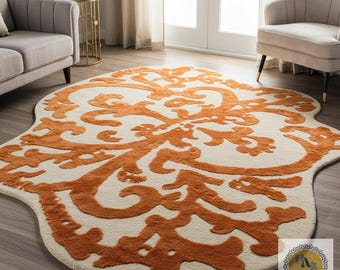 Op de afbeelding: Een ivoorkleurig vloerkleed met een groot, sierlijk, oranje bloemmotief. Het vloerkleed heeft een unieke, onregelmatige vorm en ligt op een hardhouten vloer. Het vloerkleed bevindt zich in een woonkamer met meubels.