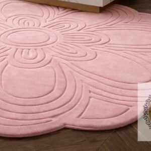 Puede incluir: Una alfombra grande y redonda de color rosa con un dise&ntilde;o floral. La alfombra presenta una forma de flor central con bordes en forma de p&eacute;talos y detalles en relieve. La alfombra est&aacute; hecha de un material suave y est&aacute; colocada sobre un suelo de madera oscura.