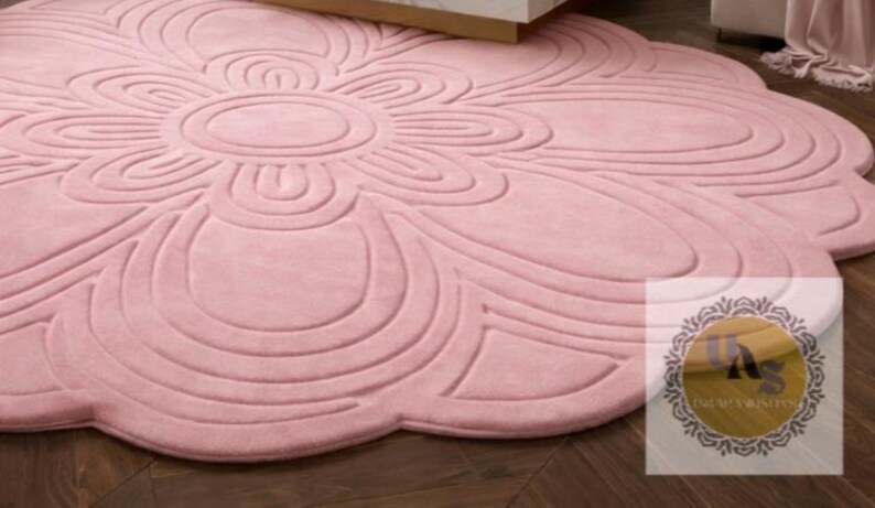 Puede incluir: Una alfombra grande y redonda de color rosa con un dise&ntilde;o floral. La alfombra presenta una forma de flor central con bordes en forma de p&eacute;talos y detalles en relieve. La alfombra est&aacute; hecha de un material suave y est&aacute; colocada sobre un suelo de madera oscura.