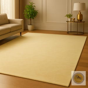 May include: A large, rectangular, solid-colored rug in a soft yellow hue. The rug is placed on a hardwood floor, with a sofa, plants, and a side table with a lamp in the background. The rug adds warmth to the room.