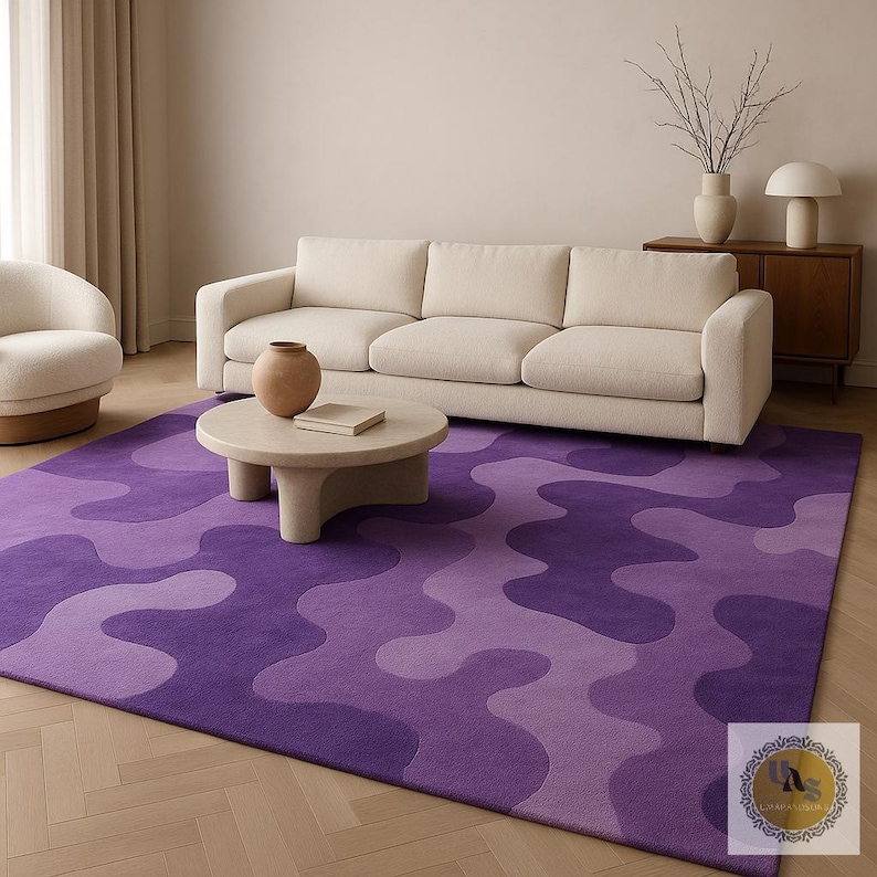 May include: A large, rectangular area rug with a wavy, abstract pattern in shades of purple. The rug is placed in a living room setting, with a cream-colored sofa and a round coffee table. The rug's design adds a modern touch to the room.