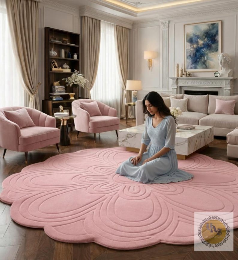 Puede incluir: Una gran alfombra rosa con forma de flor domina el primer plano de un sal&oacute;n. El dise&ntilde;o de la alfombra presenta una serie de l&iacute;neas curvas conc&eacute;ntricas. La habitaci&oacute;n incluye sillones rosas, un sof&aacute; y una mesa de centro de m&aacute;rmol. Una persona est&aacute; sentada en la alfombra.