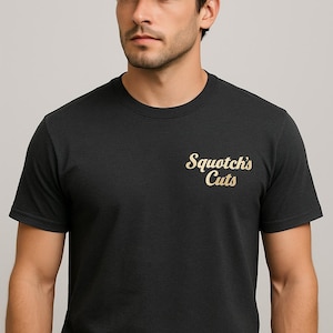 Op de afbeelding: Een zwart t-shirt met de tekst "Squotch's Cuts" in gouden letters. De persoon op de afbeelding draagt een zwarte baseball cap. Het shirt heeft een ronde hals.