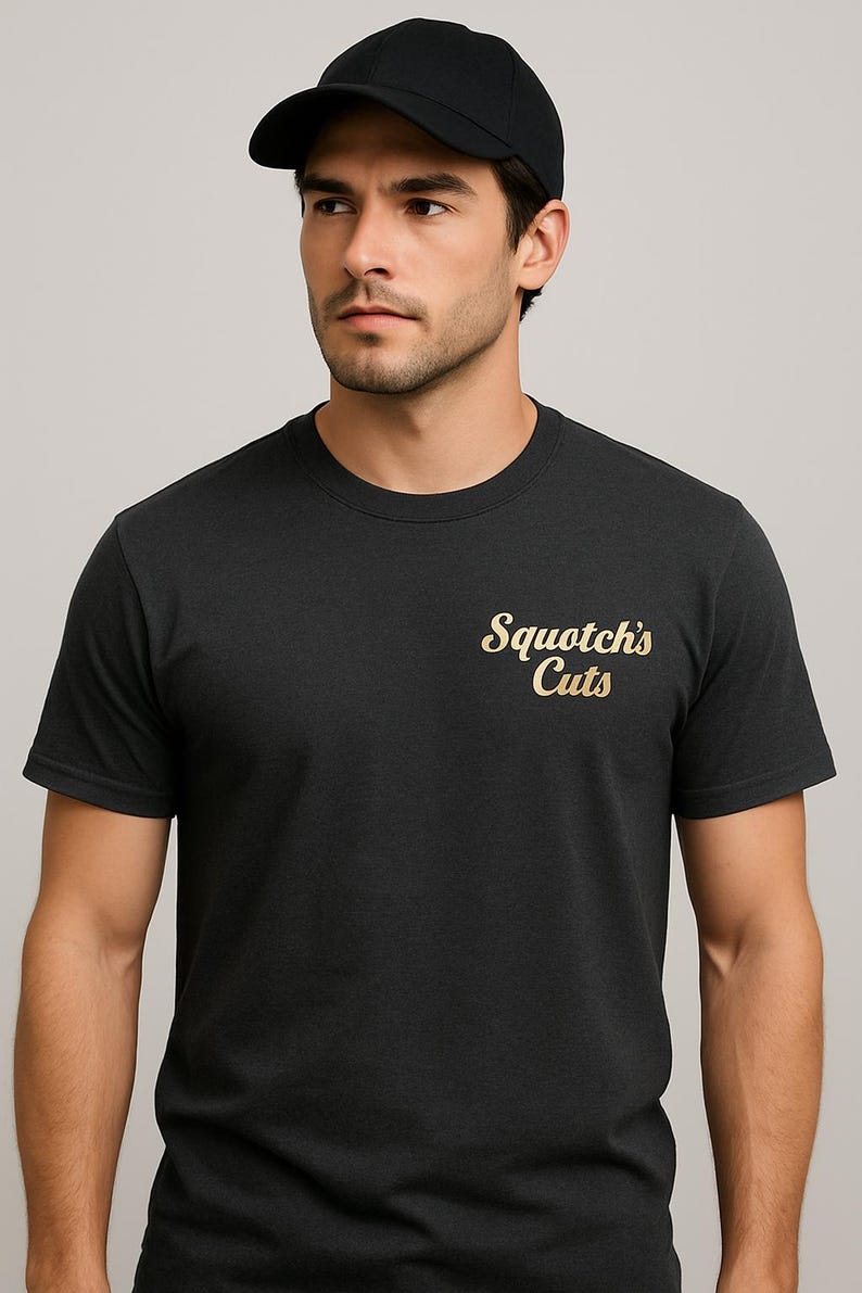 Op de afbeelding: Een zwart t-shirt met de tekst "Squotch's Cuts" in gouden letters. De persoon op de afbeelding draagt een zwarte baseball cap. Het shirt heeft een ronde hals.