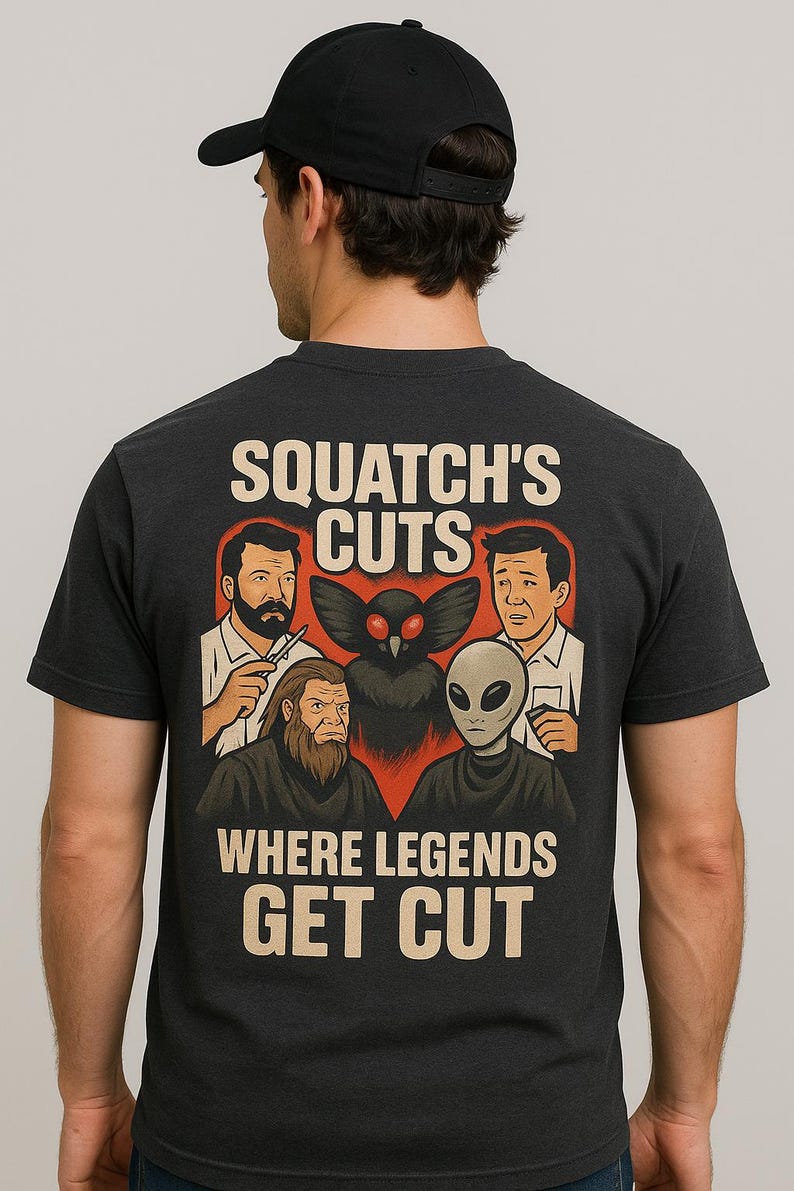 Op de afbeelding: Donkergrijs t-shirt met de tekst "SQUATCH'S CUTS WHERE LEGENDS GET CUT". De afbeelding toont cartoon illustraties van een kapper, Bigfoot, een Mothman en een alien. De persoon op de afbeelding draagt een zwarte baseball cap.