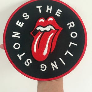 May include: Circular black patch with a red trim, displaying the Rolling Stones' famous tongue and lips emblem. The words "THE ROLLING STONES" are embroidered in white around the logo. The patch is held up by a hand.