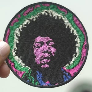 May include: Circular embroidered patch depicting a musician with black hair and a purple face. The portrait is framed by a green, white, and pink border. The patch is black and has a detailed design.