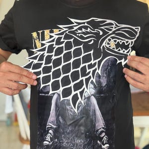 Op de afbeelding: Zwart T-shirt met een groot wit en zwart wolfskopontwerp en het woord "NE" in gouden letters. Het shirt heeft ook een grijswaardenafbeelding van een zittende persoon.