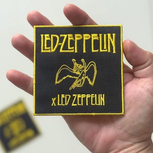 May include: A black square patch with yellow embroidered text and design. The text reads "LED-ZEPPELIN" at the top and "x LED ZEPPELIN" at the bottom. The design in the centre is a winged figure. The patch has a yellow border.