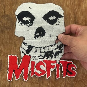 Könnte beinhalten: Gestickter Aufnäher mit dem Misfits-Schädellogo. Der Schädel ist weiß mit schwarzen Details, und das Wort "MISFITS" ist in roten Buchstaben gehalten. Der Aufnäher ist für Kleidung oder Accessoires gedacht.